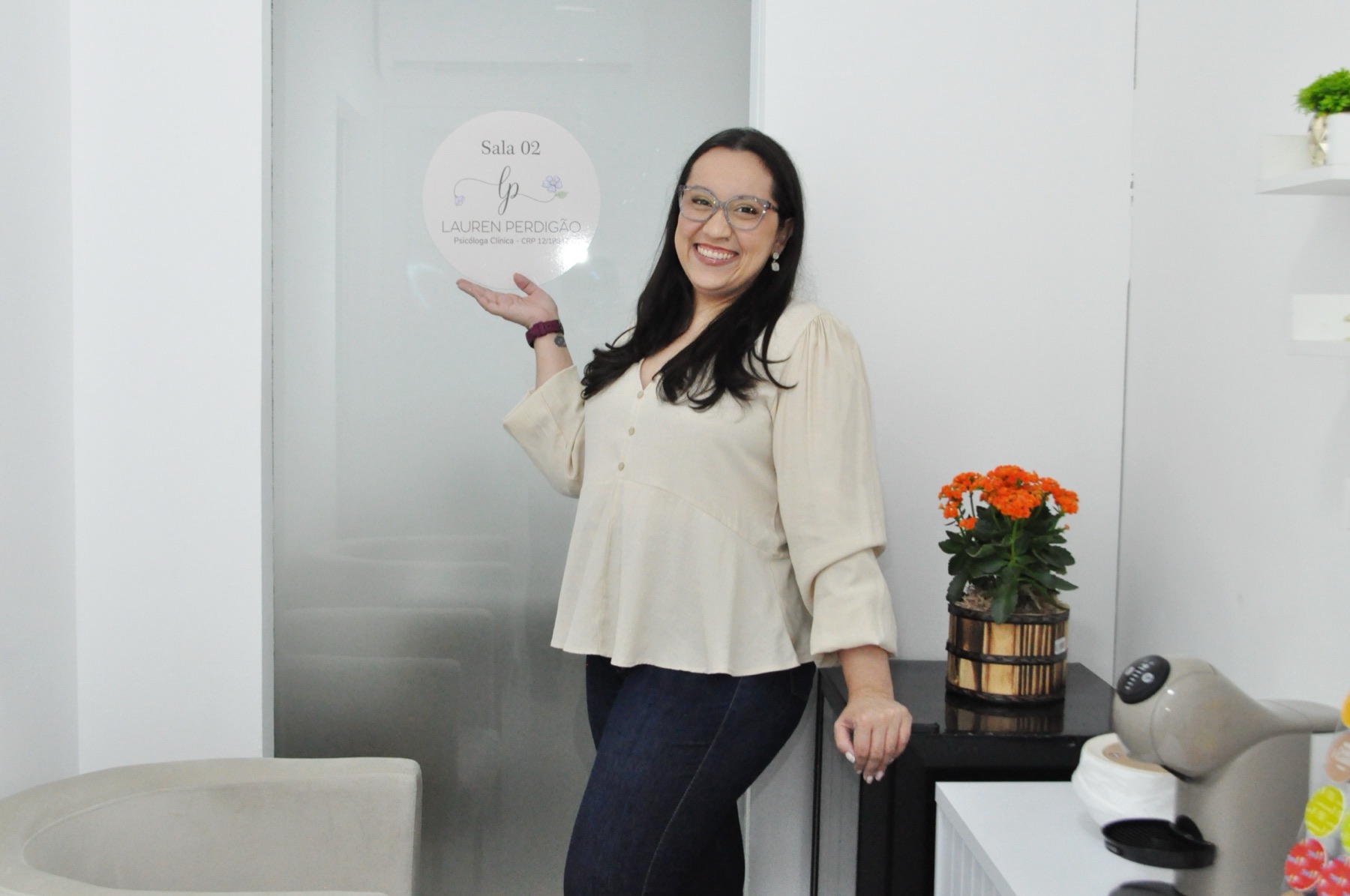 Lauren Perdigão sorrindo em seu consultório, ao lado da placa com o logo Lauren Perdigão, Sala 02
